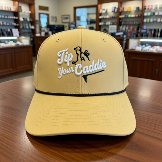Gold cap with 'Tip Your Caddie' embroidered on a wooden table in a golf pro shop